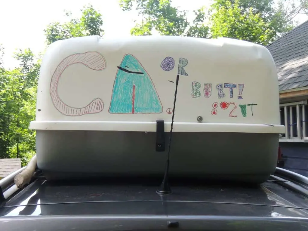 A cargo box with the words "California or Bust" on top of a small gray car.
