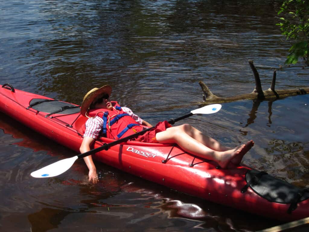 A young boy lays in a kayak on the water in the Saint Regis Canoe Area. He is wearing sunglasses and appears to be sleeping.