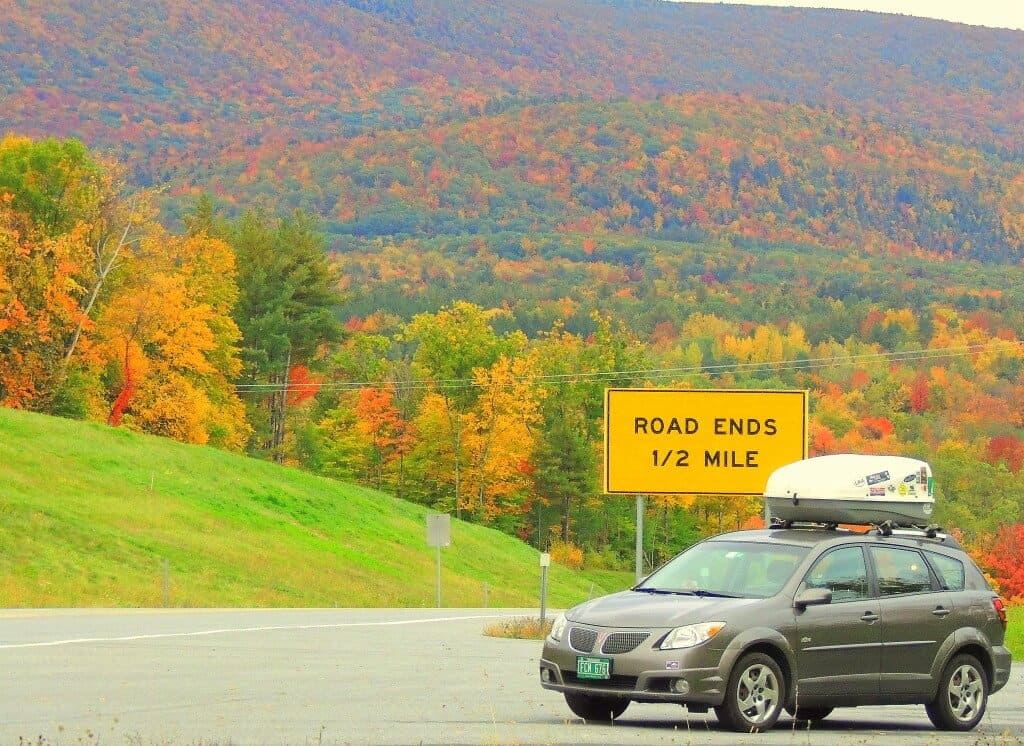 A grey compact car parked near a sign that says Road Ends 1/2 mile. Beautiful forests of fall foliage in the background