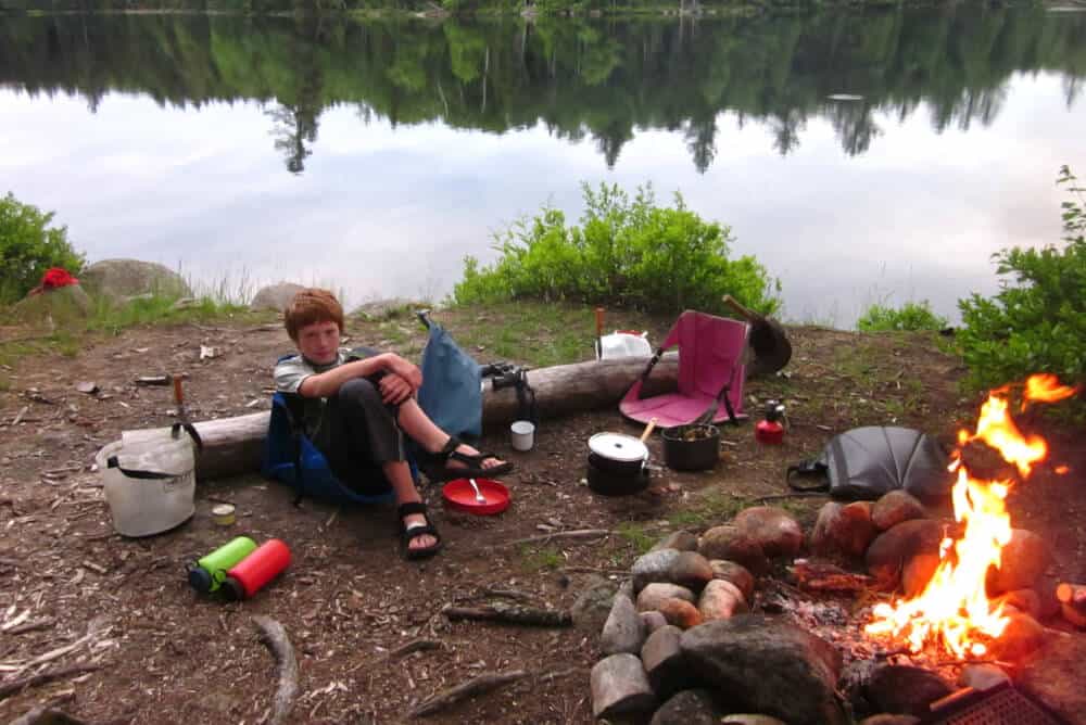 A young boy sits near a campfire. There is a lake in the background. 