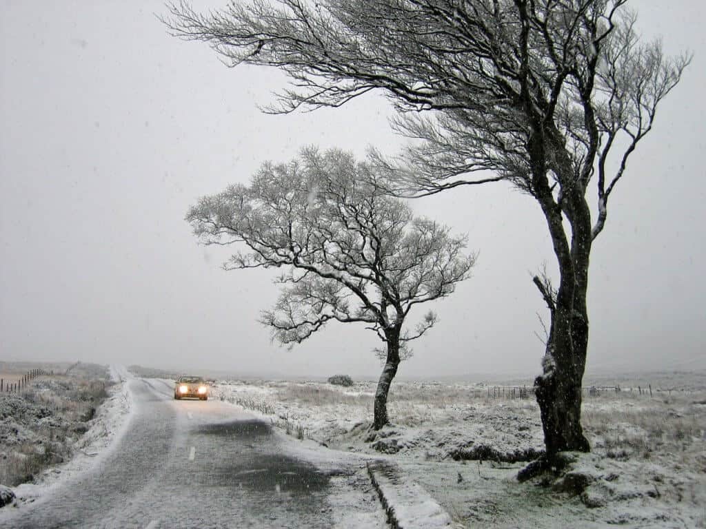 A car drives along a snowy, icy road past two bare trees.