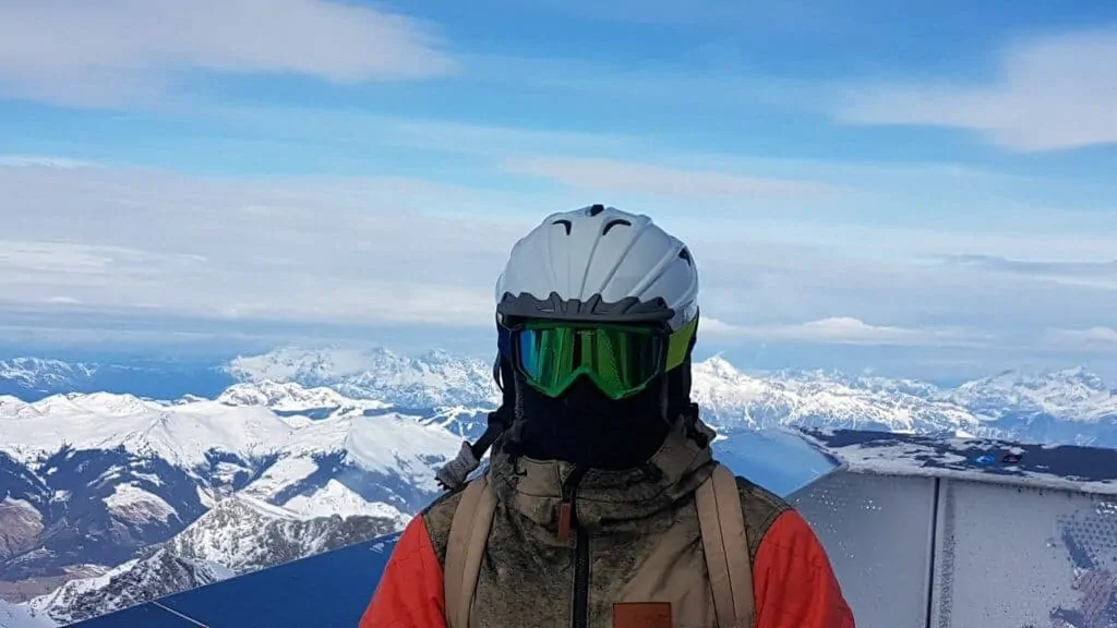 A person all decked out in ski gear stands at the top of a mountain with snow-capped peaks in the background. 