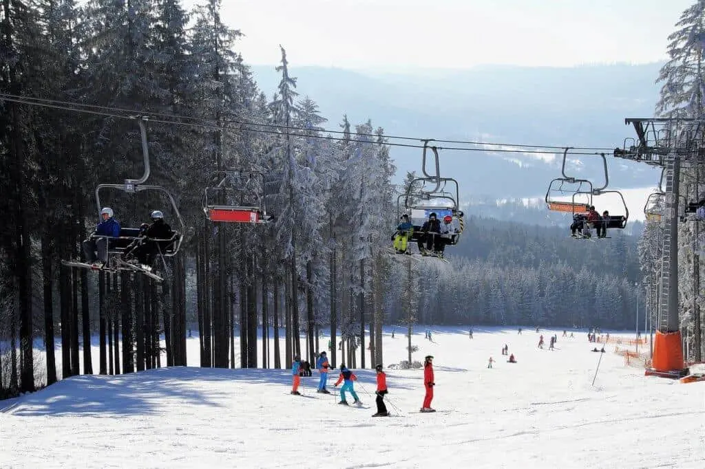A ski lift with people sitting on the chairs. Underneath there are a lot of skiers wearing colorful ski jackets. 