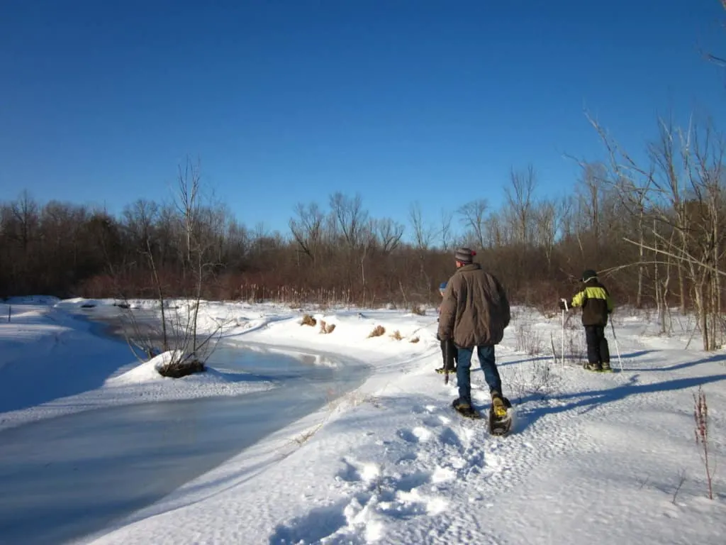 A man and two kids walk on snowshoes near a frozen pond.