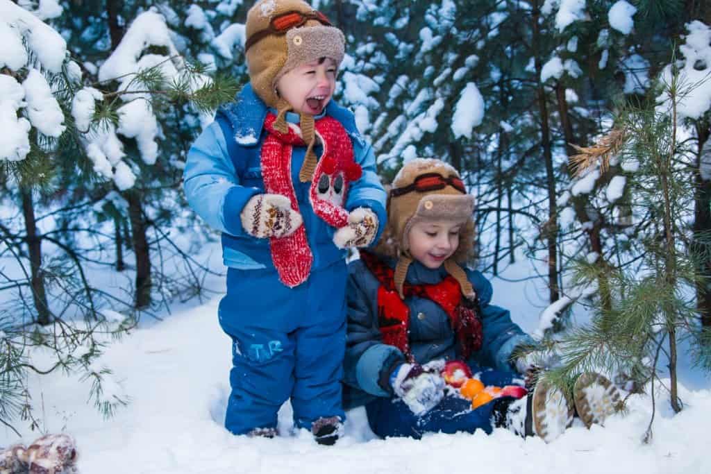 Two boys in winter clothing stand in a snowy forest. One of the boys is holding some oranges. 