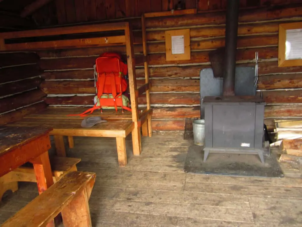 The inside of a New England camping cabin at Merck Forest. Picture shows a woodstove, some wooden bunks, and an orange backpack.