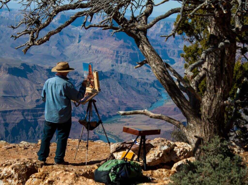 A man painting on an easel at the edge of the Grand Canyon. 