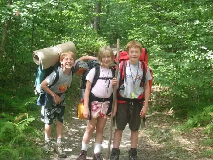 Three kids wearing backpacking packs standing on a trail after a long hike.