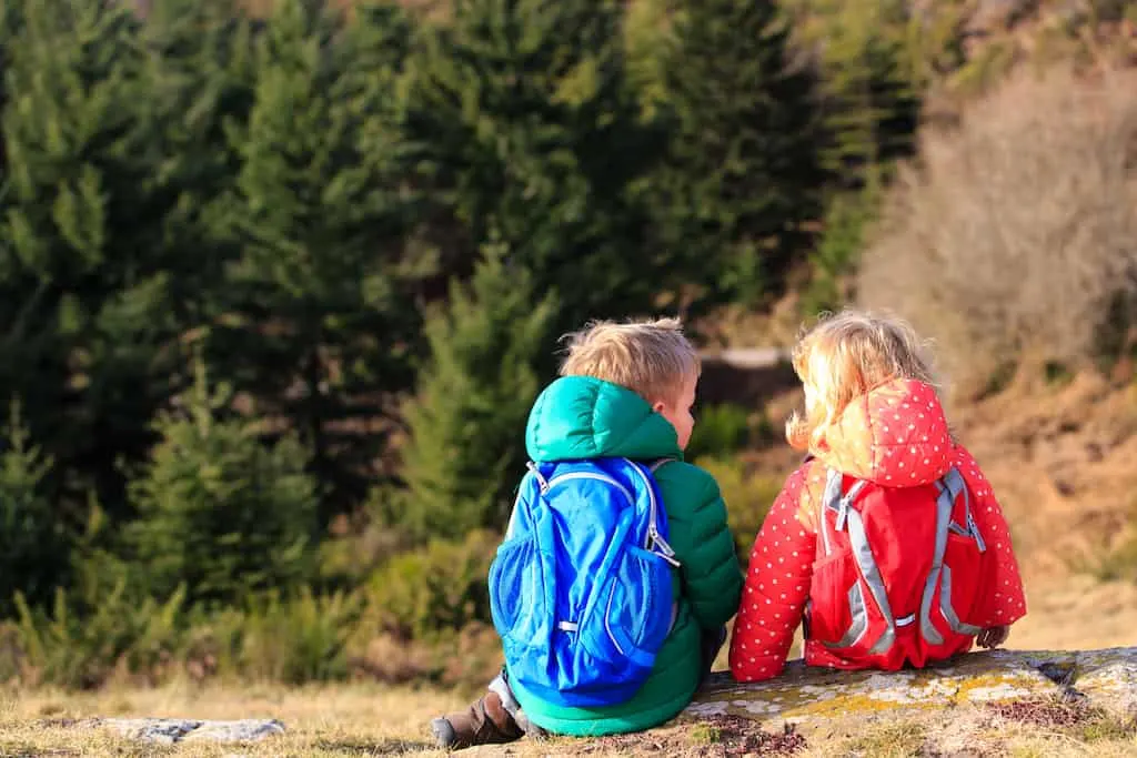 Two kids sit with their backs to the camera. They are wearing colorful clothing and hiking daypacks.
