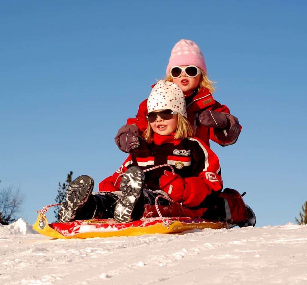 Two girls in winter clothes sit on a sled at the top of a hill.