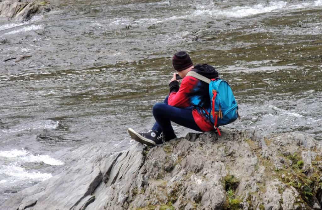 A boy sits on a rock near a stream in the winter. He is wearing colorful clothing and a blue daypack.