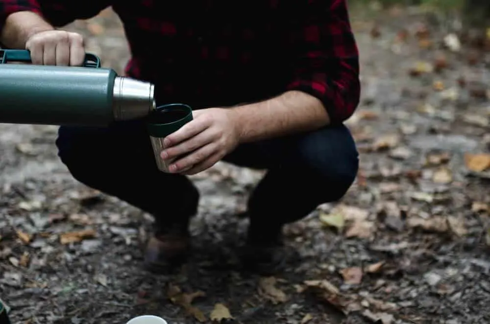 A man kneels outside pouring hot chocolate from a thermos into a mug.