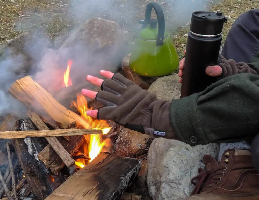 a close up of a campfire with hands warming up and a tea kettle.