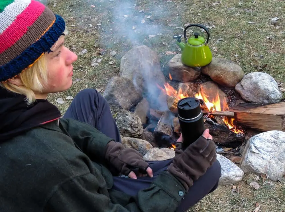 A young man sits near a campfire enjoying a hot drink. 