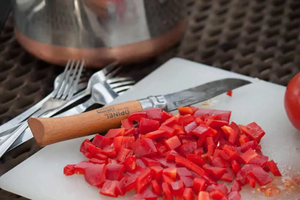 A cutting board with chopped red peppers on it, next to a folding pocket knife and four forks. 