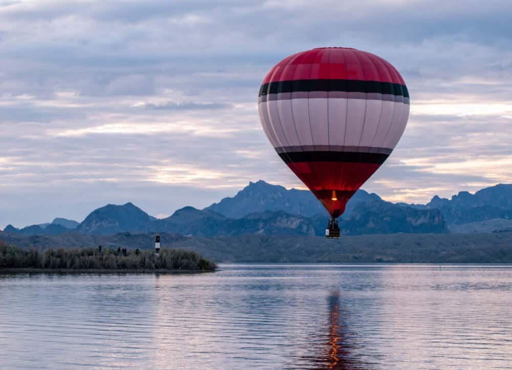 A red and white striped hot air balloon floating near the surface of Lake Havasu with mountains in the background.
