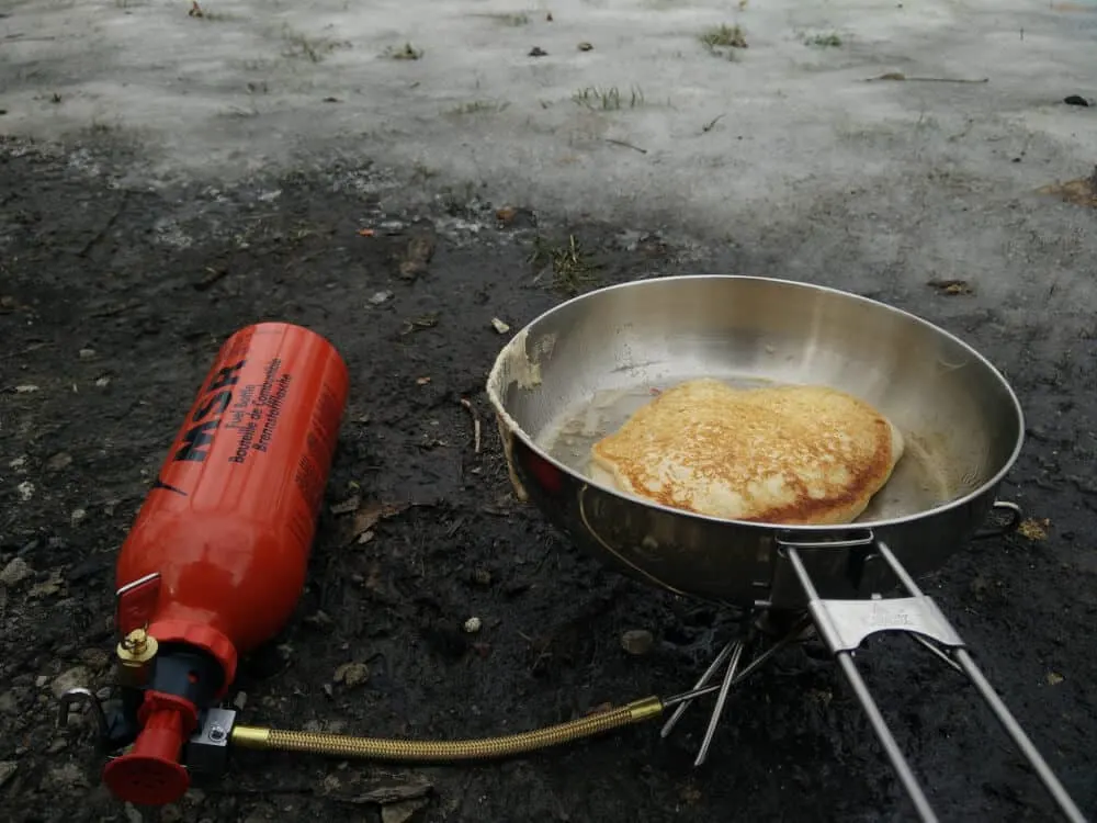 A pancake cooks in a frying pan over a small camping stove.