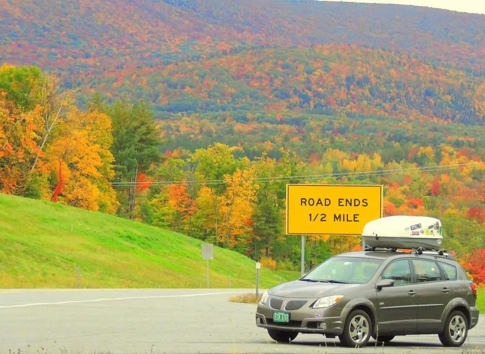 A grey Pontiac Vibe with a cargo carrier on top. Fall foliage in the background and a yellow sign that says, 