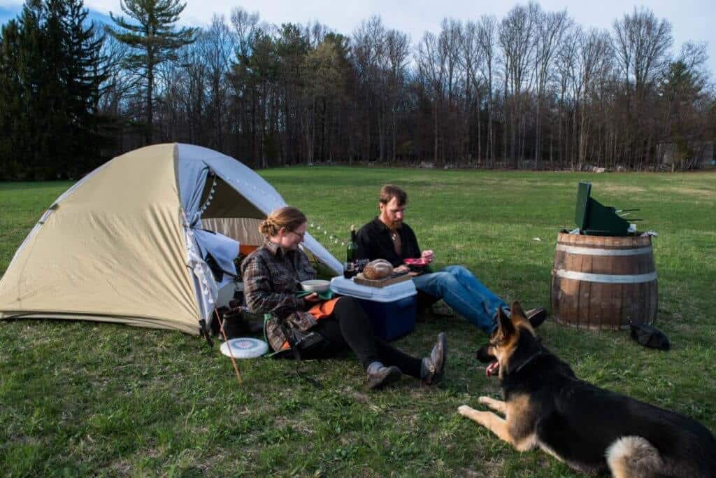 camping etiquette two people and a German Shepherd sitting in front of a small tent eating a meal.
