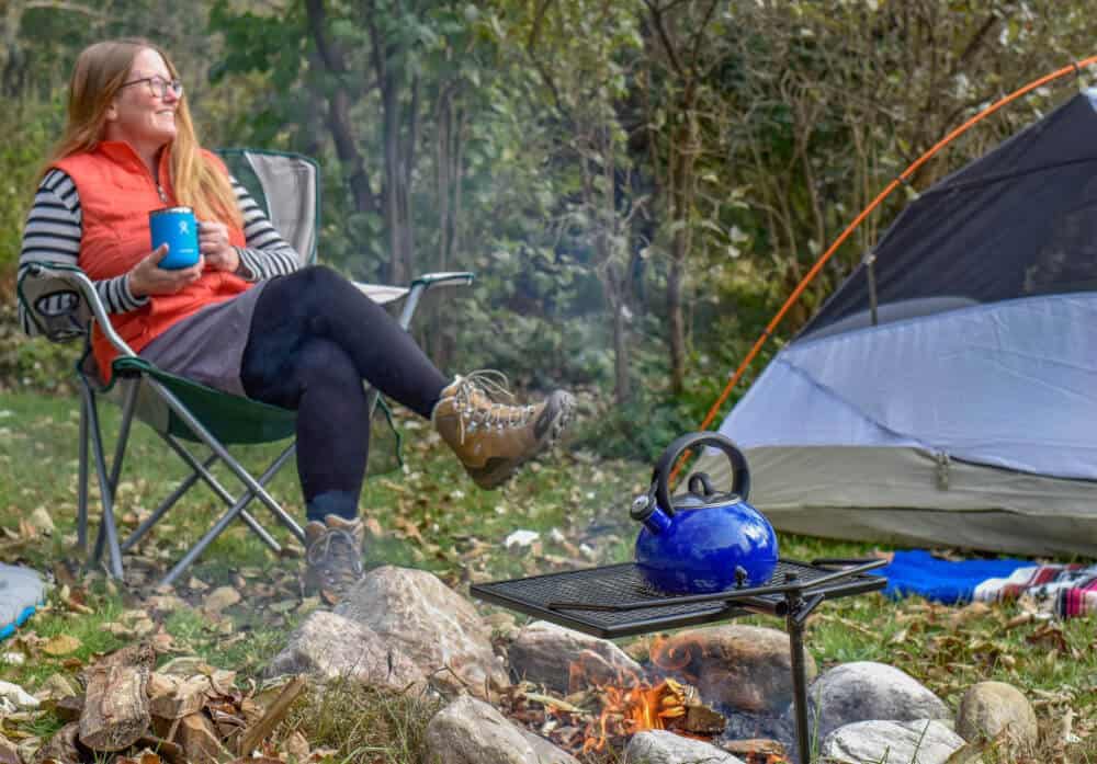 A woman sits near a camp fire holding a cup of steaming tea.