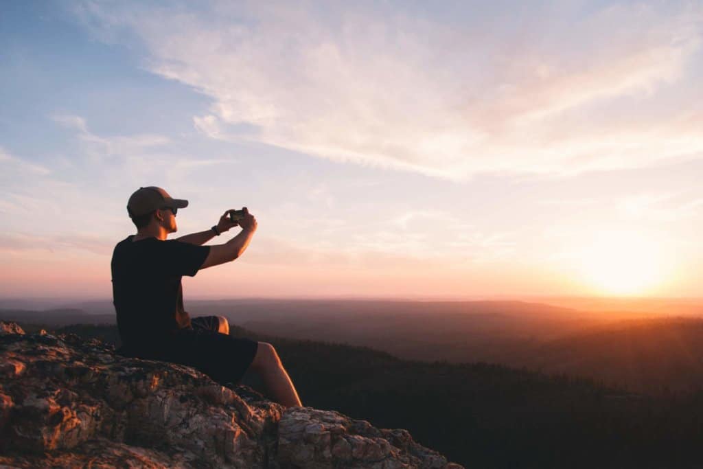 A man sits on a rock facing the setting sun holding a smartphone.