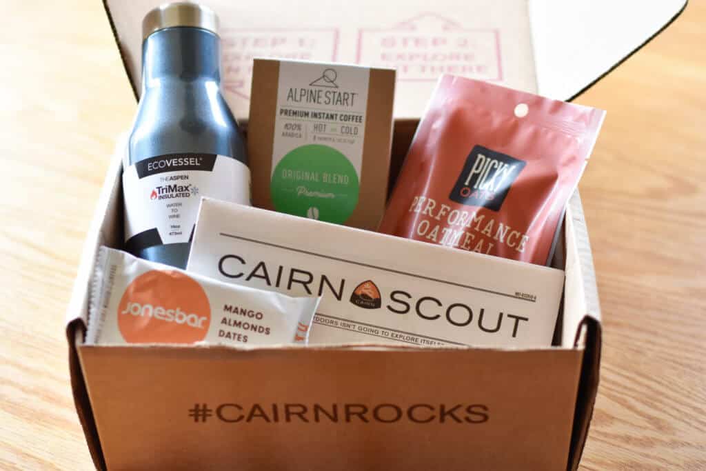 A recent Cairn subscription box, featuring oatmeal, a granola bar, instant coffee, and a water bottle. They are all arranged in a cardboard box.