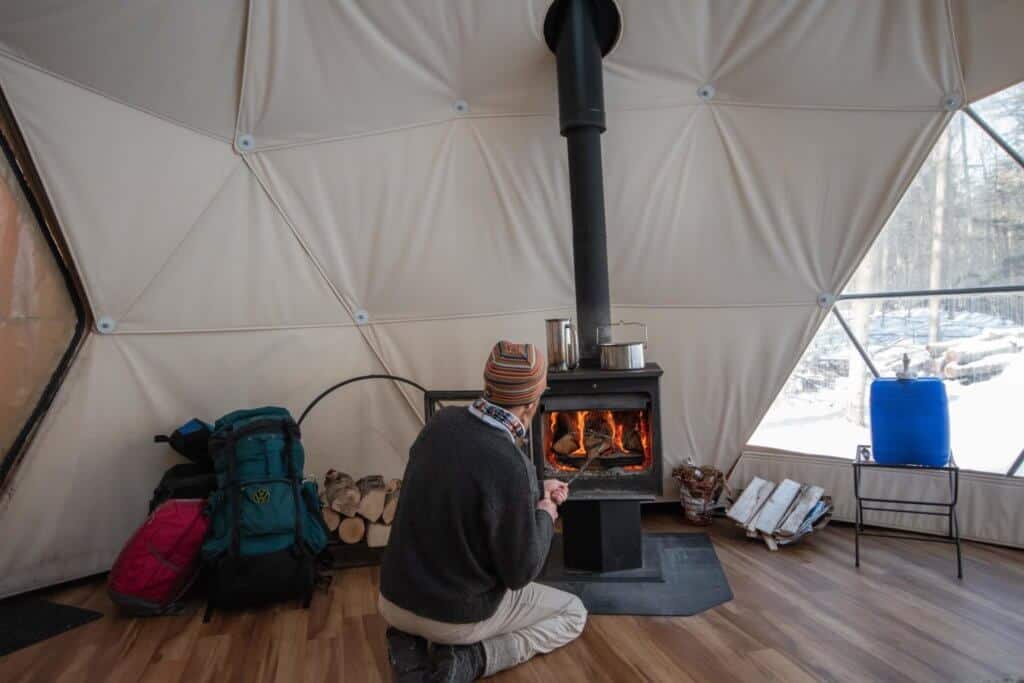 A man kneels down in front of a woodstove inside a geodesic dome in Putney, Vermont.