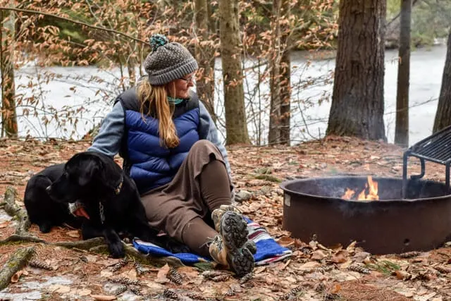 A woman sits near a campfire with a black Labrador.