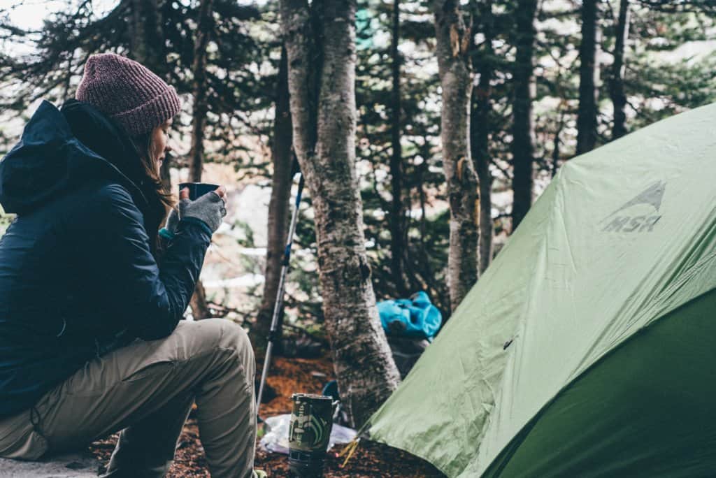 A woman sits near a tent holding a warm mug and wearing winter clothing.