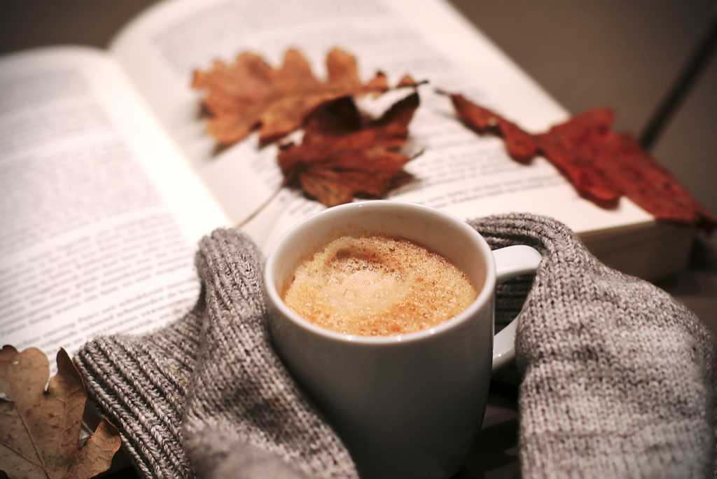 An open book with fall leaves, a coffee cup, and the sleeves of a wool sweater.