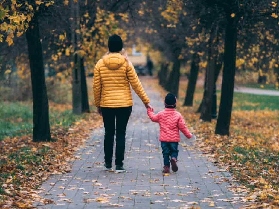 A woman and a little girl are seen from the back walking down a brick path covered with fall leaves.