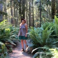 A woman stands in the middle of a hiking trail next to tall ferns and very tall trees.