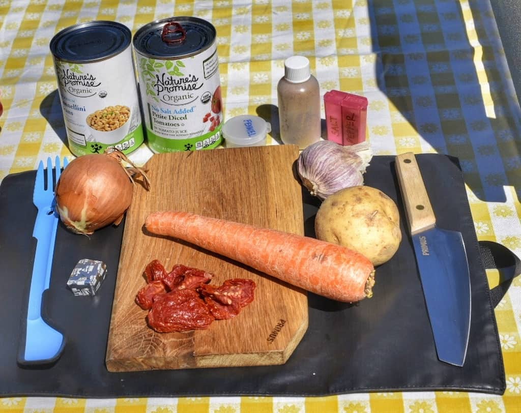 Various ingredients needed to make white bean tomato sou when camping. There are veggies on a cutting board, several cans of beans and tomatoes, and a chef's knife.