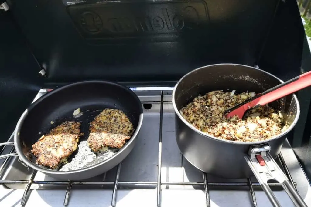 A camping stove with a pot and a frying pan on it. The pan has two vegetarian quinoa patties cooking and the pot contains the quinoa patty batter. 