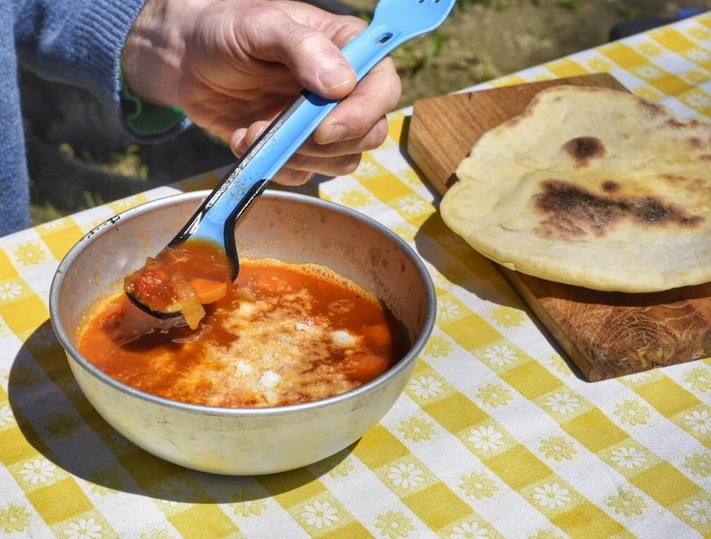 A bowl of white bean tomato soup next to a piece of flatbread on a yellow and white checked tablecloth