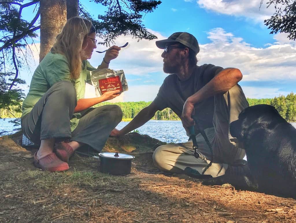 Two people sit on the shores of a lake sharing a backpacking meal by Trailtopia
