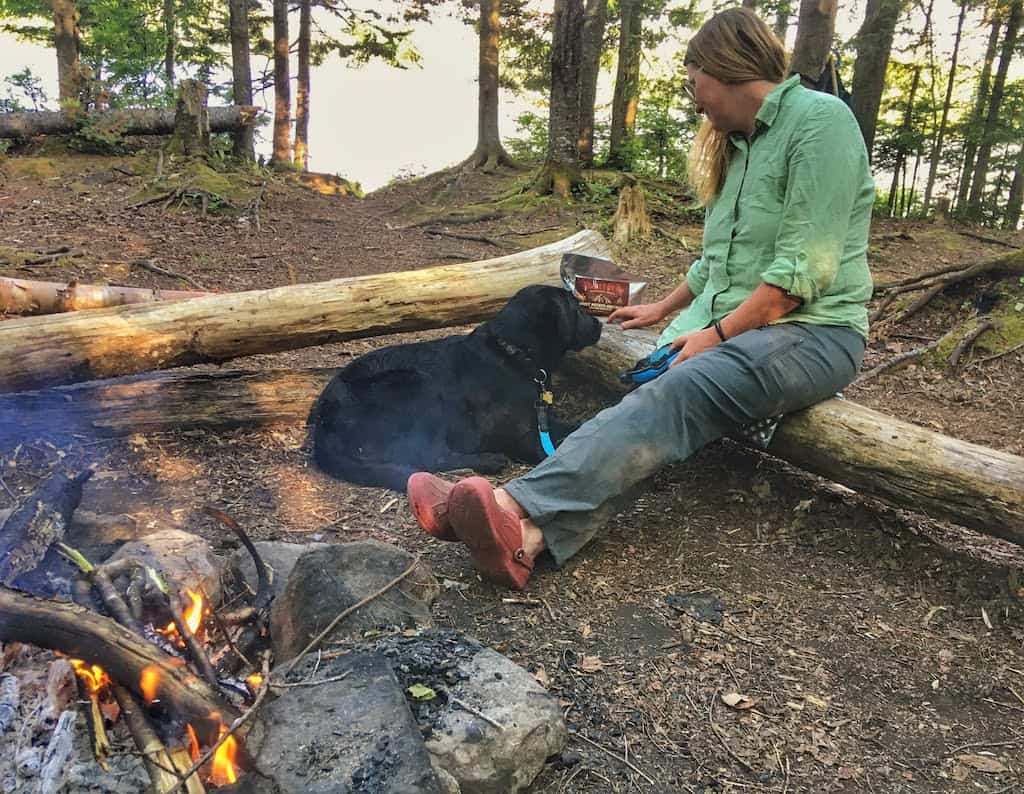 A woman sits near a campfire with a black Labrador Retriever