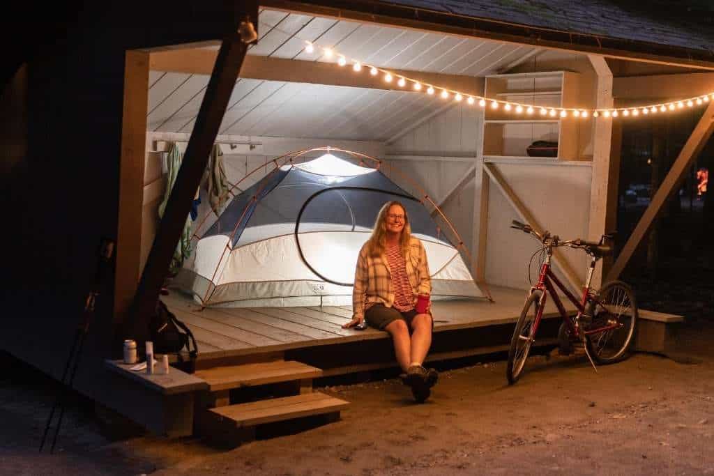 A woman sits in a lean-to next to a lit-up tent at night. There is a bike next to her and twinkly lights hanging from the ceiling. Jamaica State Park, Vermont.