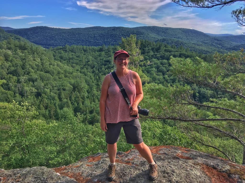 A woman stands at the top of Little Ball Mountain in Jamaica State Park Vermont. She is wearing shorts and a t-shirt and has a camera strapped around her neck.