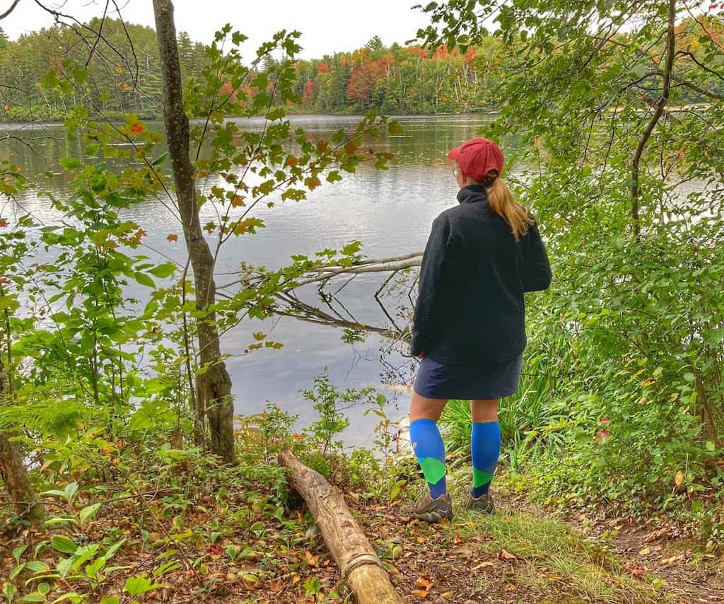 A woman stands on the shore of a lake looking out at the water. She is wearing Lily Trotters compression socks. 