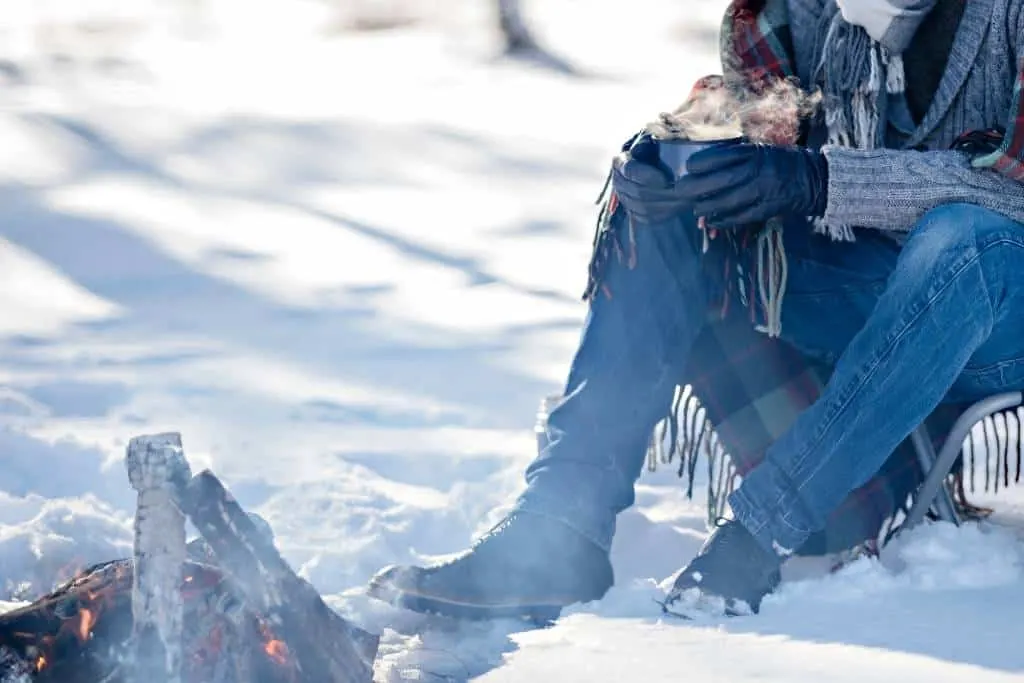 a person sits in front of a campfire in the snow enjoying a hot drink.
