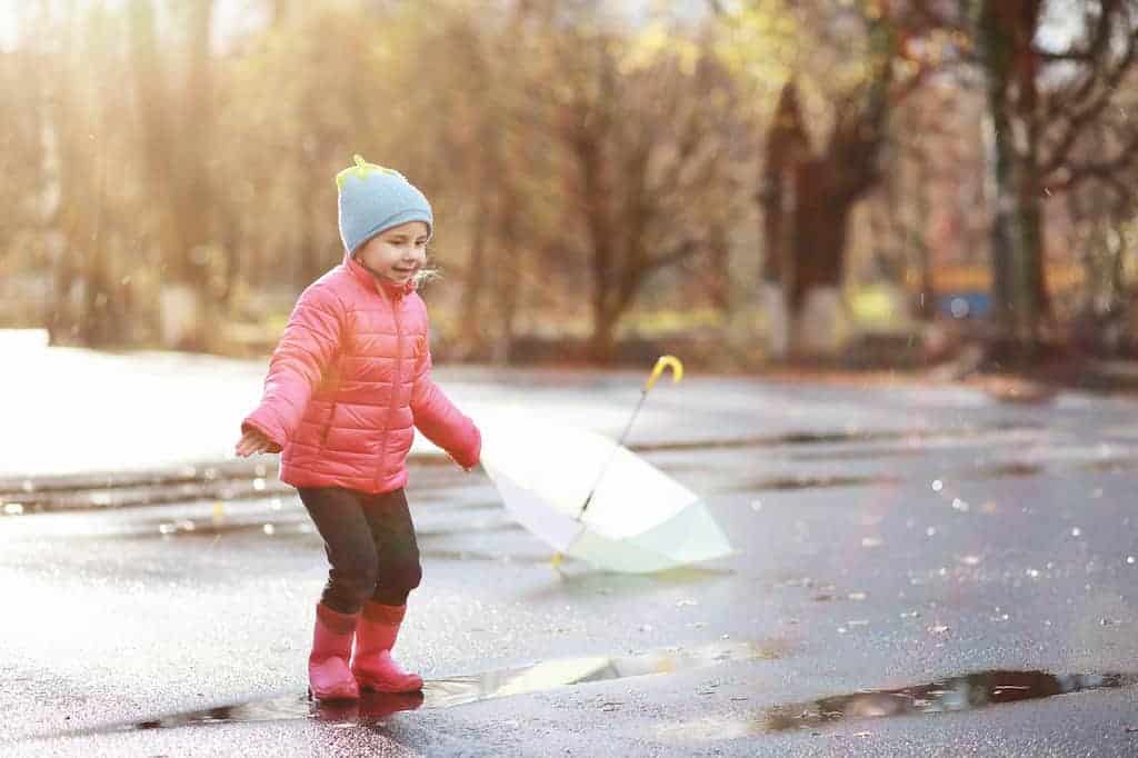 A child stands in a mud puddle wearing warm winter clothes for toddlers. She has a smile on her face.