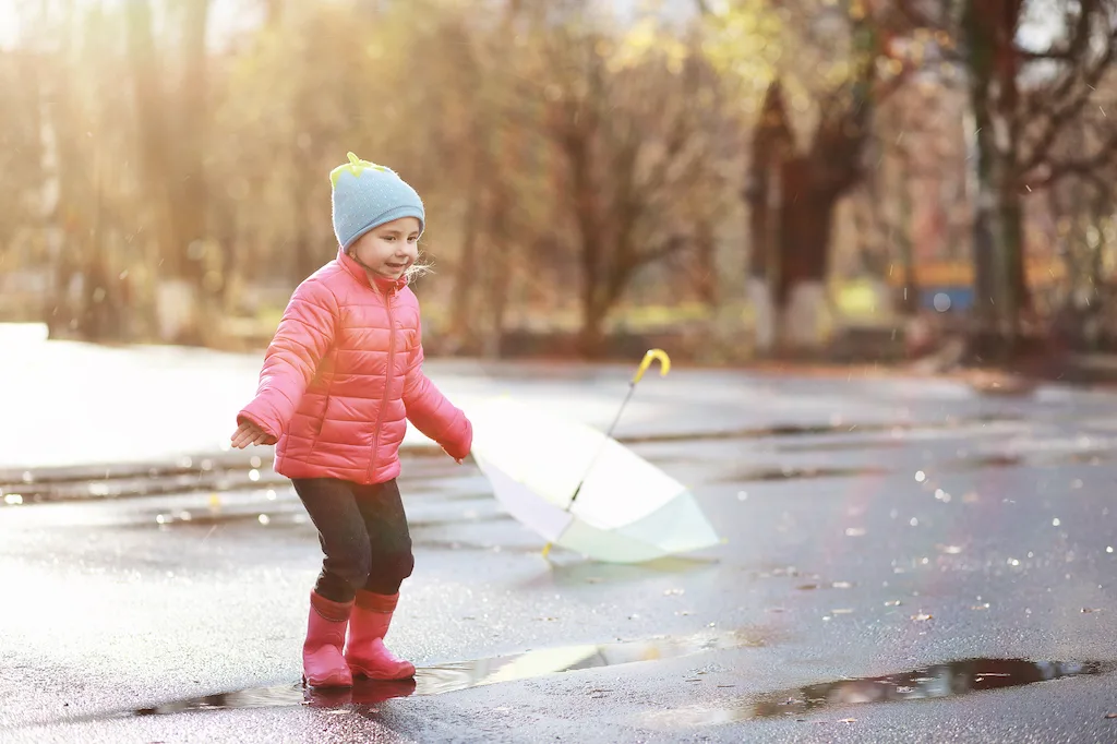 A child stands in a mud puddle wearing warm winter clothes for toddlers. She has a smile on her face.