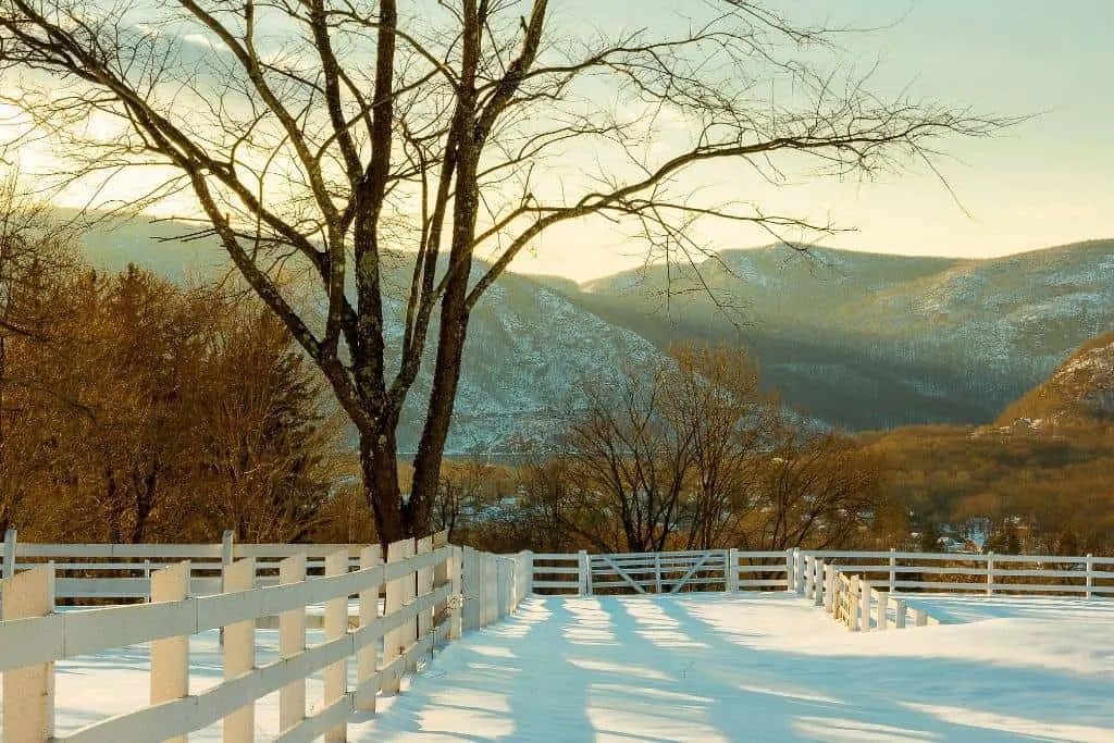A view of a Hudson Valley, New York farm field with a white fence around it covered with snow. There are mountains in the background. 