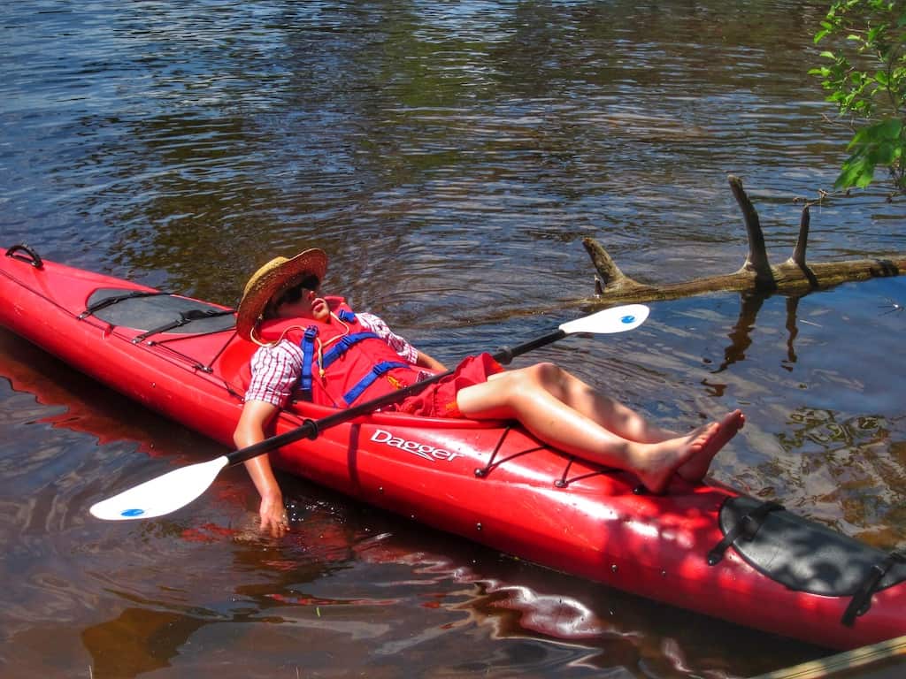 A young boy lounges in a red kayak on a pond. He is wearing a wide-brimmed hat, a life jacket, and sunglasses.