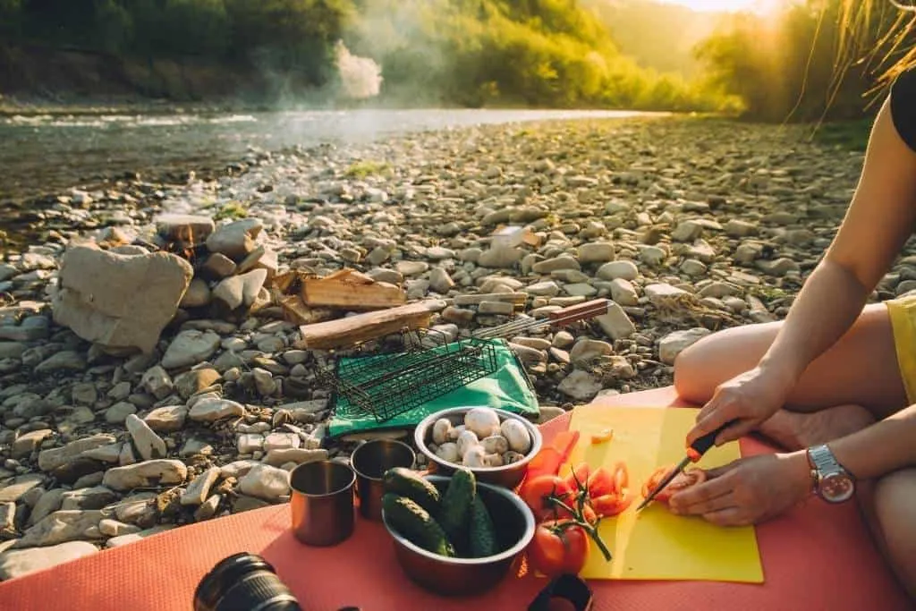 A woman sits on a camp pad slicing tomatoes near a campfire. 