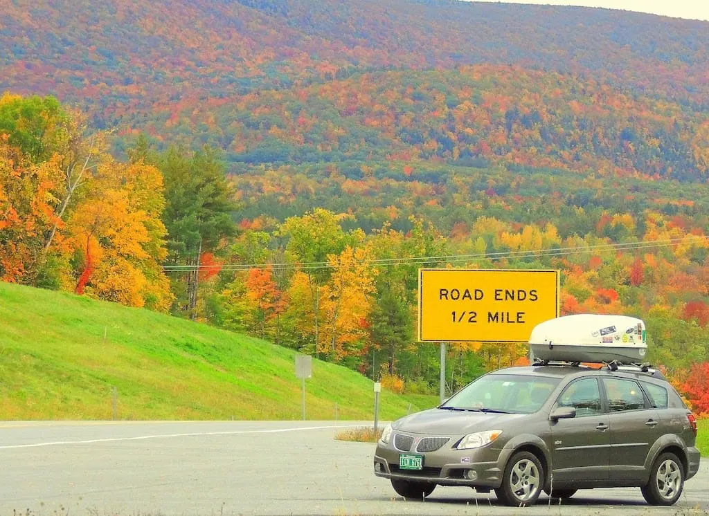An image of a small car in front of a sign that says "road ends 1/2 mile". There is lush fall foliage in the background.