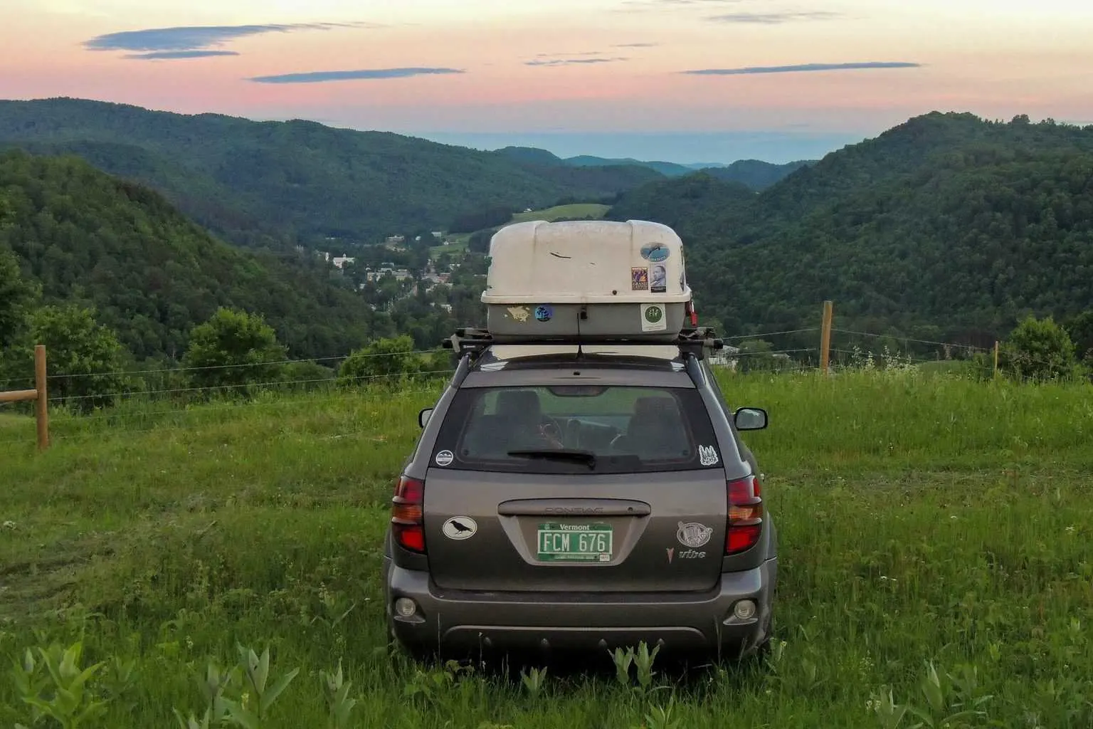 The back of a grey car with a white cargo carrier on top of it pointing toward a mountain sunset.