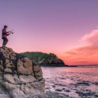 a person stands on a large rock to watch the sunset over the Bay of Fundy.