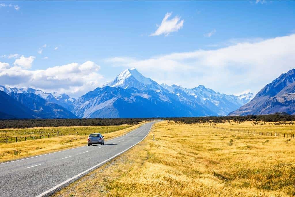 A small car drives on a desolate stretch of road toward huge snowcapped mountains.